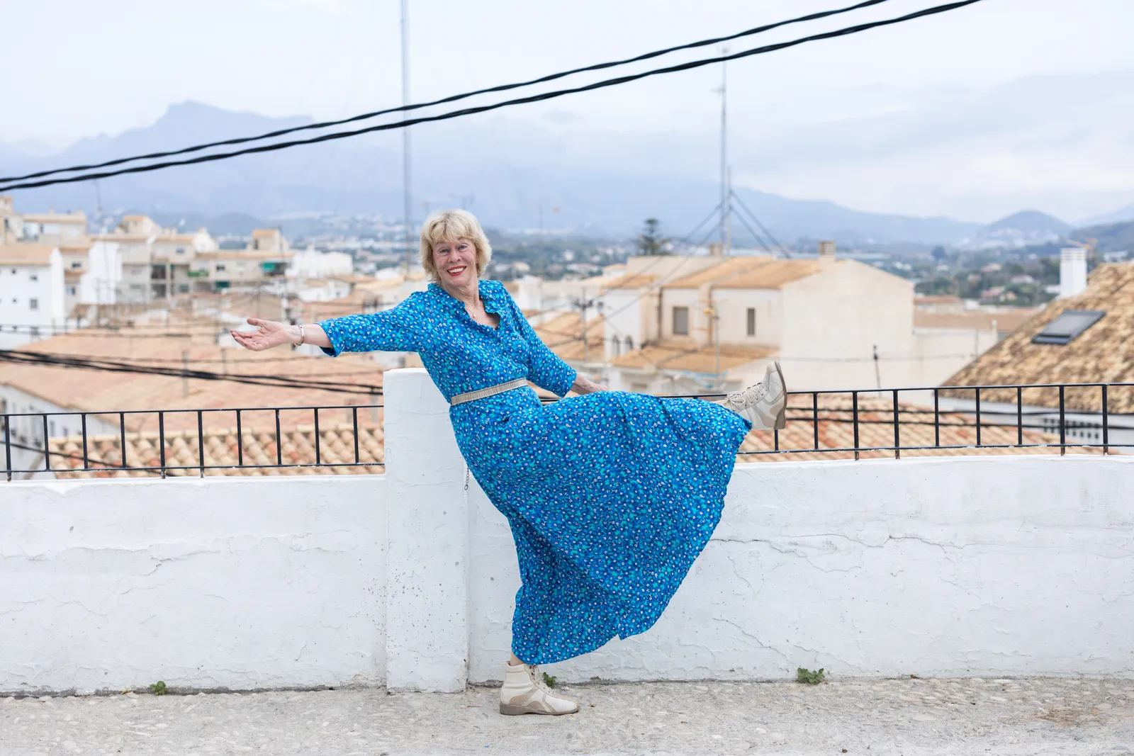 Jessica Larive in een lange blauwe jurk zwaait lachend met een been omhoog bij een wit muurtje boven de daken
