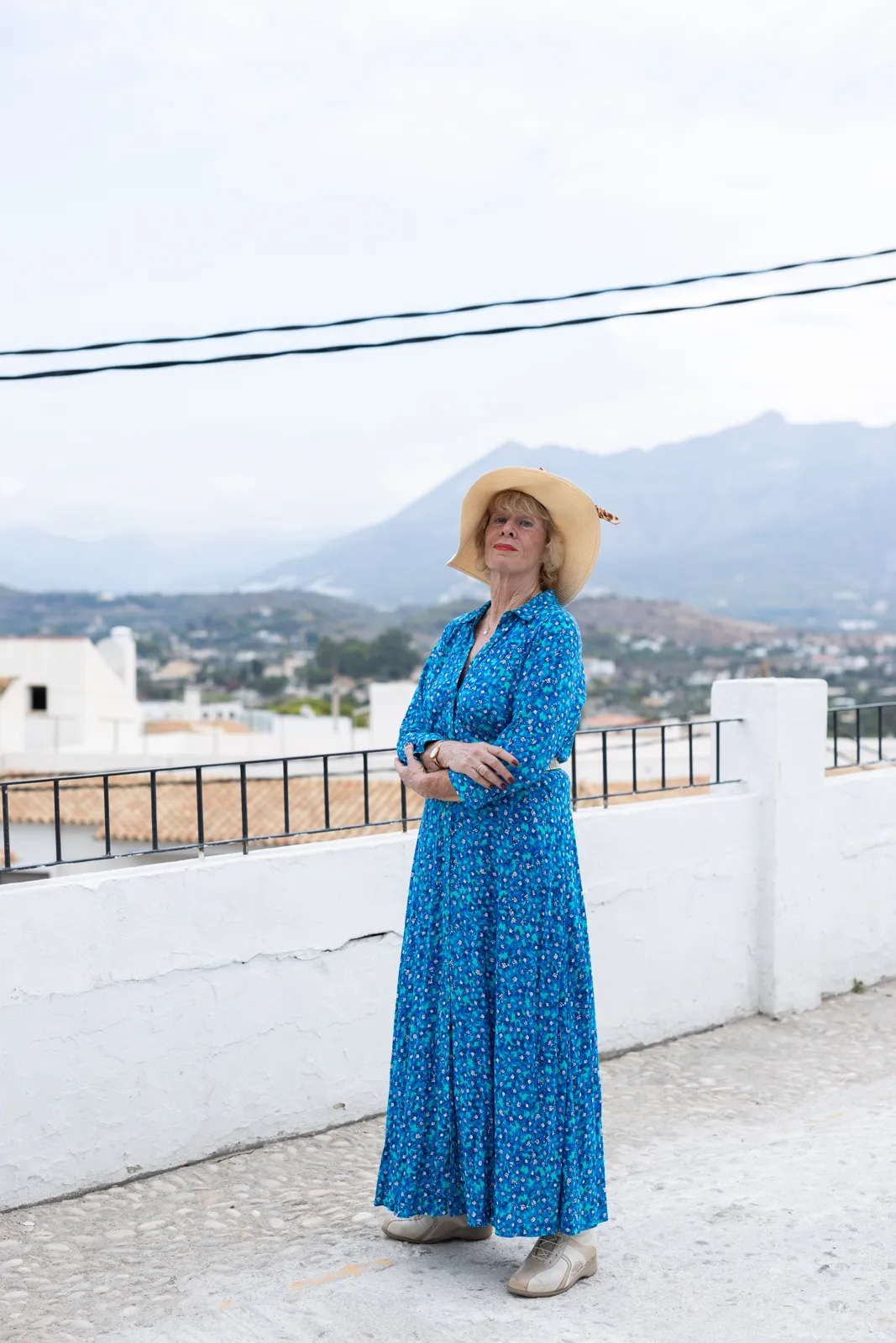 Jessica Larive in een lange blauwe jurk en met een strohoed staat met gekruiste armen op een wit terras, met bergen op de achtergrond