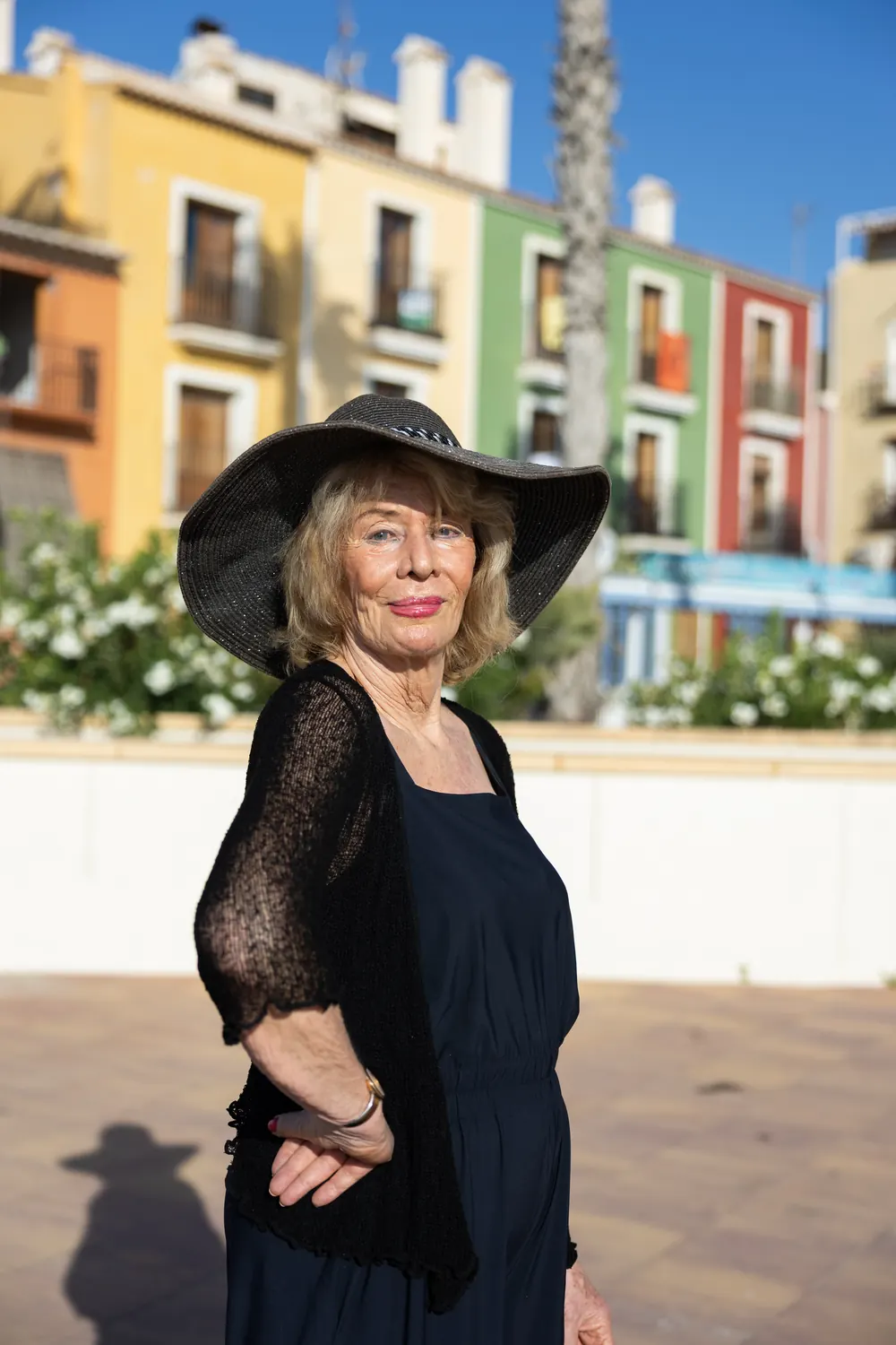 Jessica Larive in het zwart met een grote zwarte hoed, voor kleurige huizen