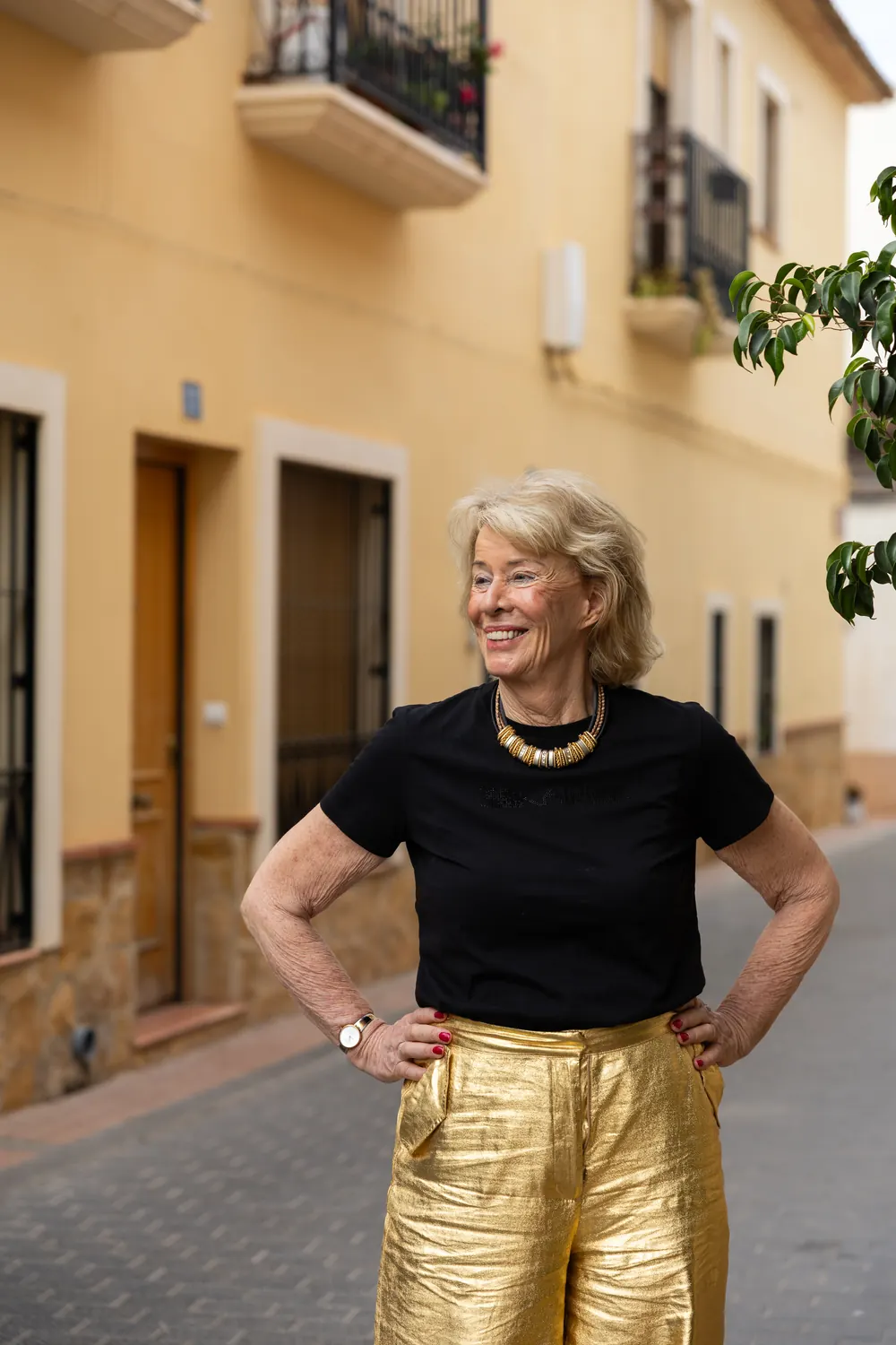Jessica Larive lacht in een smalle straat, in een zwart shirt en een gouden broek, met haar handen in de zij